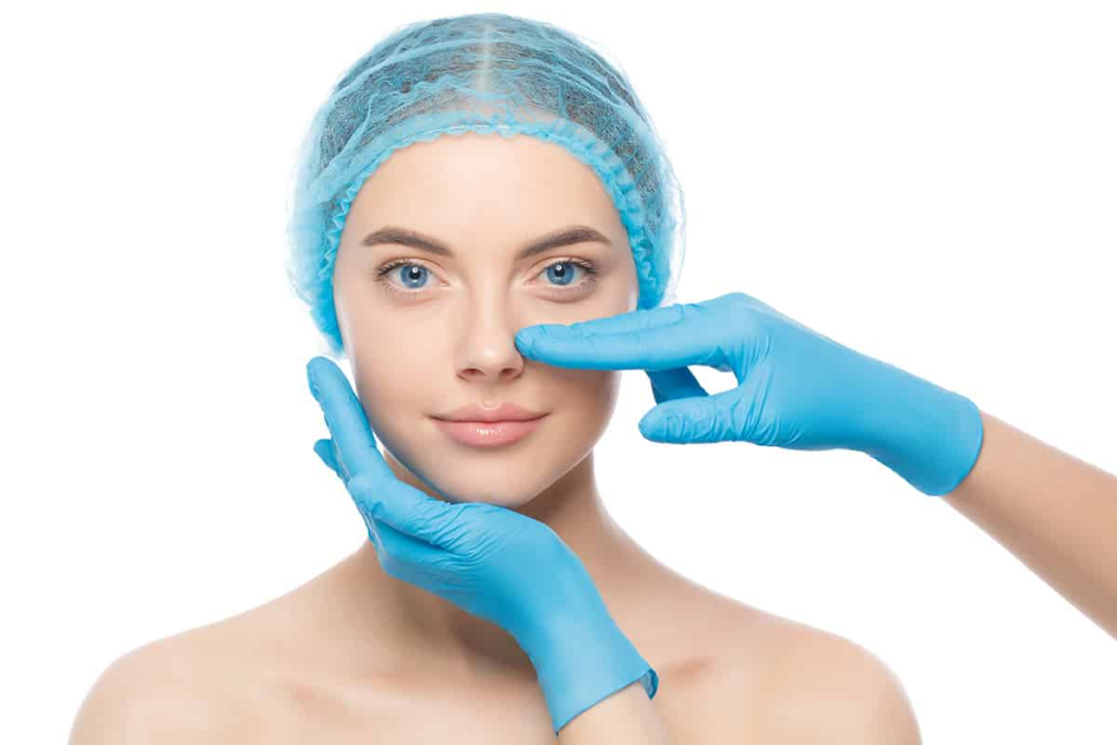A woman wearing a surgical cap and gloves is pointing to her nose, likely in a medical or cosmetic procedure context.