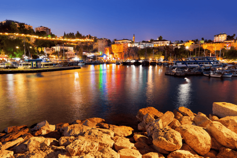 A serene nighttime view of a harbor with boats docked along the waterfront, illuminated by warm lights reflecting on the calm water.