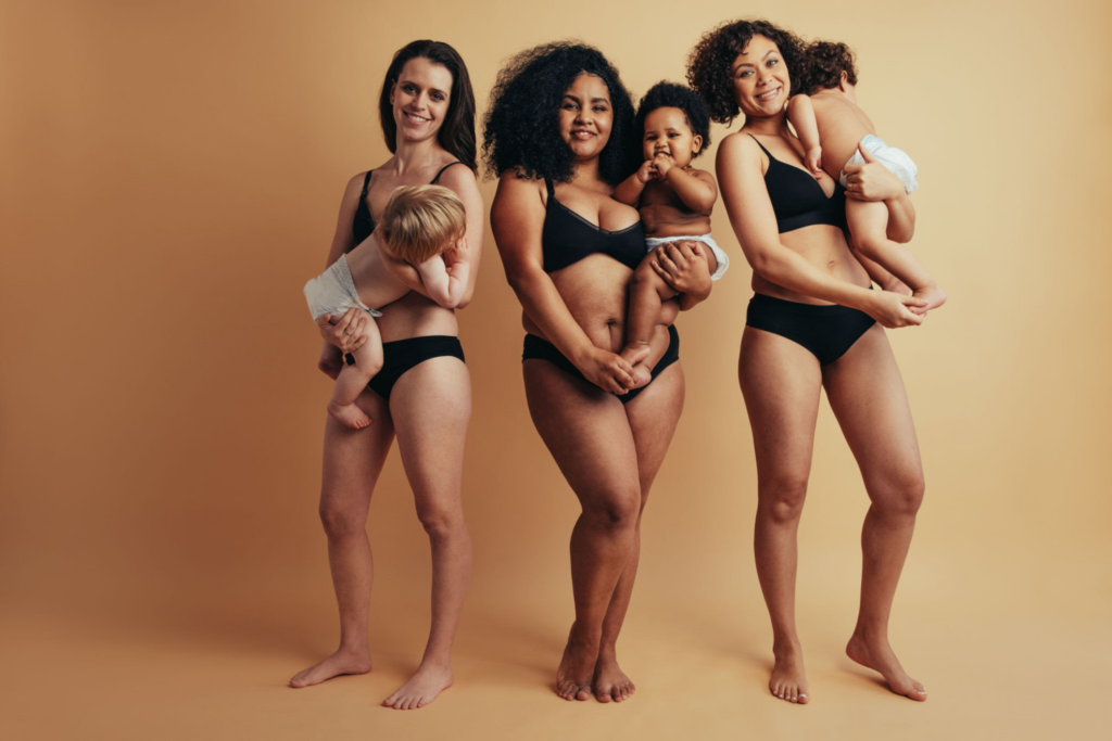 Four women standing together, each holding a baby. They are all wearing black bikini bottoms and are barefoot. The background is a solid peach color.