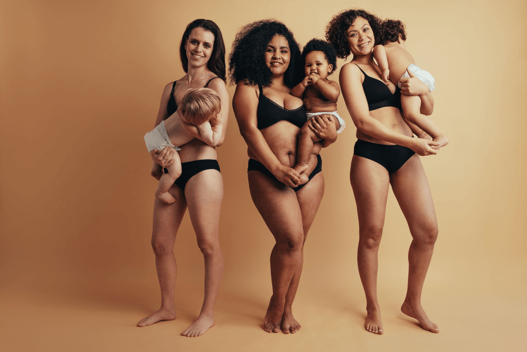 Four women standing together, each holding a baby. They are all wearing black bikini bottoms and are barefoot. The background is a solid peach color.
