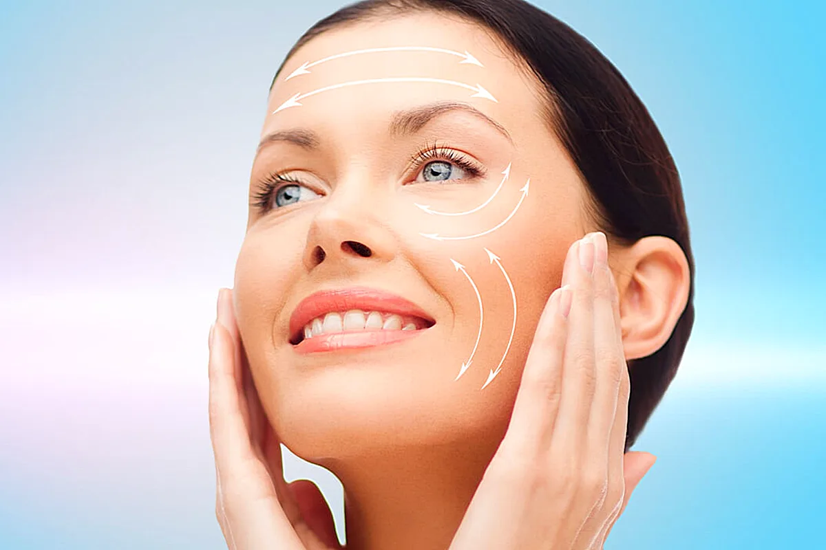 Una donna è mostrata con frecce che indicano le aree del suo viso, probabilmente evidenziando le regioni per una procedura cosmetica o medica. Sorride e sembra trovarsi in un ambiente clinico.