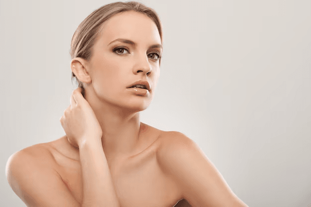 A woman with blonde hair posing against a plain background, touching her neck with her right hand.