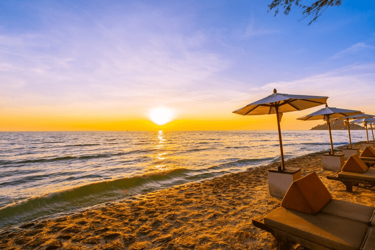 A serene beach at sunset with the sun partially visible above the horizon, casting a golden glow over the ocean. The beach has several lounge chairs and umbrellas arranged along the shore.