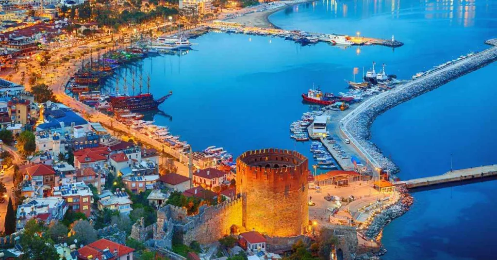 Aerial view of a coastal city with a prominent castle by the harbor, illuminated against the night sky. The harbor is filled with boats and yachts, and the cityscape includes brightly lit buildings along the waterfront.