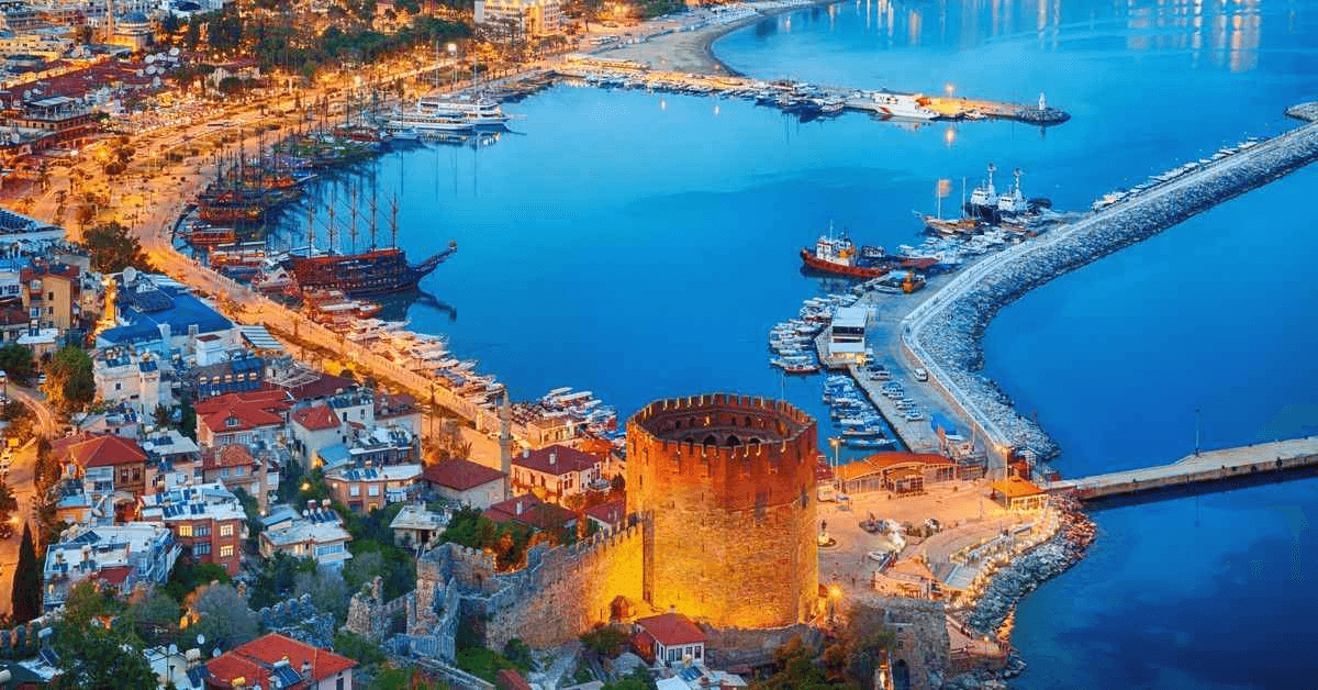Aerial view of a coastal city with a prominent castle by the harbor, illuminated against the night sky. The harbor is filled with boats and yachts, and the cityscape includes brightly lit buildings along the waterfront.