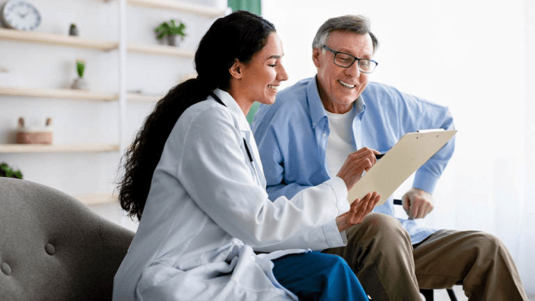 A doctor and an elderly patient are sitting together, looking at a clipboard. The doctor is wearing a white coat and the patient is wearing a blue shirt. They are both smiling.
