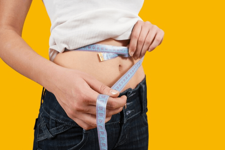 A person measuring their waist with a tape measure against a yellow background.