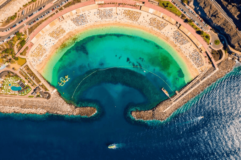 An aerial view of a large, curved lagoon with crystal-clear turquoise water, surrounded by a sandy beach with numerous sun loungers and umbrellas. The lagoon is connected to the open sea by a narrow channel, and there are boats visible in the water.
