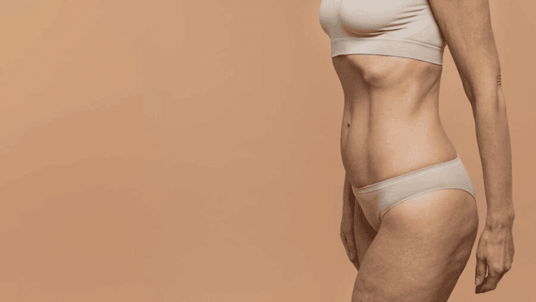 A person with a toned physique stands against a peach-colored background. The individual is wearing light-colored undergarments, showcasing well-defined abdominal muscles.