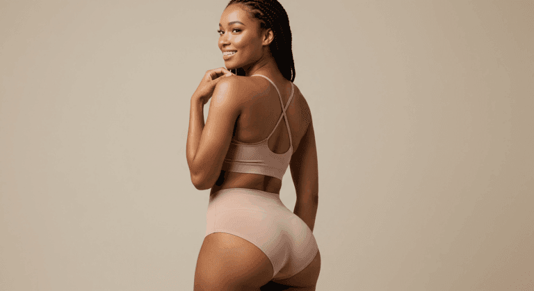 A woman wearing a beige sports bra and matching high-waisted leggings, posing with her hands on her hips, smiling over her shoulder. She has dark hair styled in braids and is standing against a plain beige background.