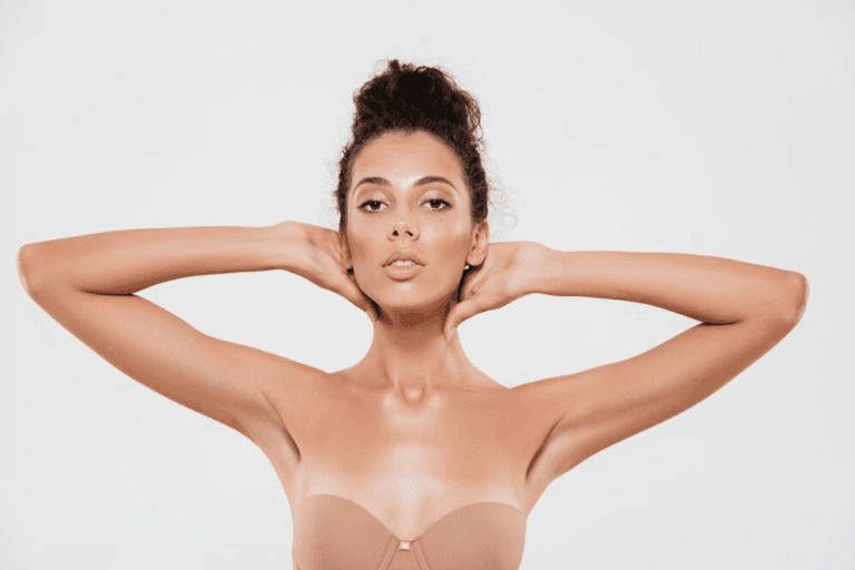 A woman with her arms behind her head, wearing a strapless top, posing confidently against a plain white background.