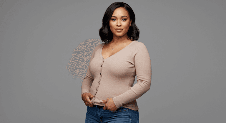 A woman with dark hair wearing a light pink sweater and blue jeans, posing against a gray background.