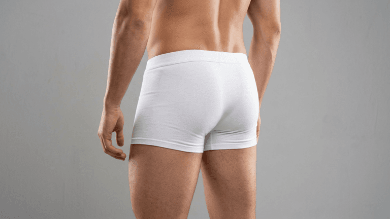 A close-up, rear-view shot of a muscular male torso and upper thighs, wearing clean white boxer briefs against a neutral gray background. The lighting is soft and even, highlighting the masculine anatomy and muscular definition of the back, shoulders, and legs. The camera is positioned at hip level, focusing on the fit of the underwear, with no visible makeup, jewelry, or distracting background elements.