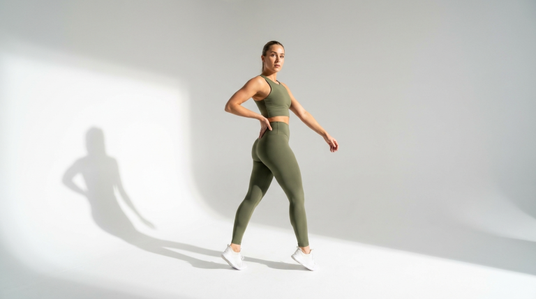 A fit, athletic woman with long brown hair tied back, wearing an olive green sports bra and matching high-waisted leggings, poses in a dynamic walking stance against a plain white studio background. The studio lighting creates soft shadows, highlighting her toned anatomy. She is wearing white athletic sneakers. The camera captures her from a full-body side-profile angle as she looks directly toward the lens. Her makeup is natural and minimal, suitable for a fitness-themed photoshoot. The overall atmosphere is clean, professional, and health-focused.