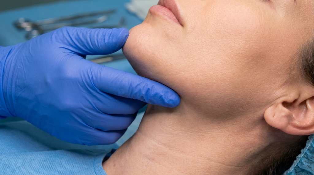 Close-up clinical shot of a female patient undergoing a pre-surgical jaw examination. The camera is positioned at a close-range angle, focusing on the jawline and neck area. The lighting is bright and clinical, characteristic of a sterile medical environment. The patient has natural skin without visible makeup, and her anatomy shows a clear, smooth jawline. She is wearing a sterile blue medical gown and a hair cap. In the background, blurred stainless steel surgical instruments are visible on a light blue sterile drape, emphasizing the professional, hygienic surgical setting, while a surgeon's hand, wearing a blue medical glove, gently palpates the patient's chin.