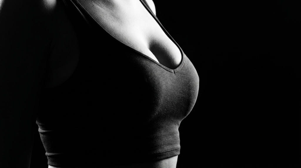 A close-up, black and white side-profile shot of a female torso, focusing on the chest area. The subject is wearing a form-fitting black sports bra. The image features dramatic chiaroscuro lighting, emphasizing the curves and anatomy of the chest against a solid, dark black background. The camera angle is at a mid-level side view, highlighting the texture of the skin and the silhouette. No makeup or facial features are visible.