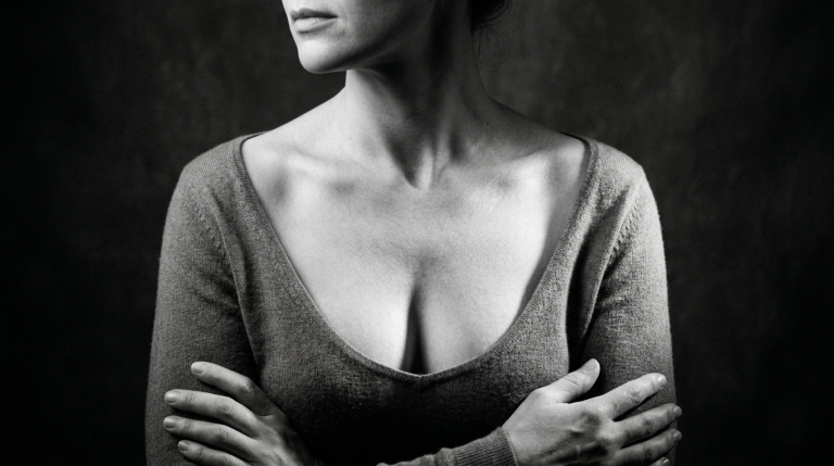 A black and white studio portrait of a woman with feminine features and natural, minimal makeup. The lighting is soft and dramatic, creating subtle highlights on her collarbone and shoulders against a dark, textured background. She has a slender build with a prominent collarbone, wearing a long-sleeved, deep v-neck knit sweater. Her arms are crossed, emphasizing her posture and neckline.