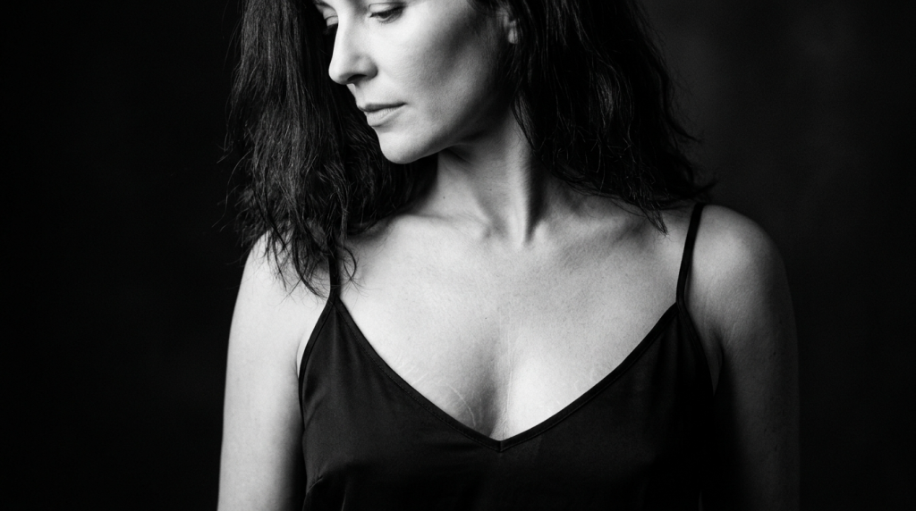 A high-contrast black and white portrait of a woman with a feminine profile, featuring soft, dramatic side-lighting that emphasizes her jawline and natural skin texture. She has subtle, minimal makeup and wavy hair. She is wearing a simple black camisole top with thin straps, revealing natural anatomical details on her chest, set against a dark, minimalist, and out-of-focus background.