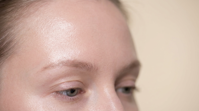A close-up, high-angle shot focusing on the forehead and eyes of a fair-skinned female individual. The lighting is soft and bright, highlighting a natural, dewy skin texture without visible makeup. The person has light-colored eyes and natural, neatly shaped eyebrows. The background is a clean, neutral, and out-of-focus beige surface.