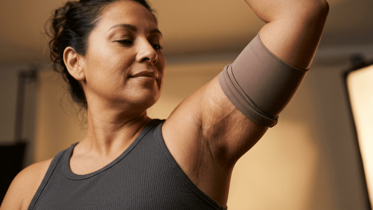 A medium close-up shot from a low angle shows a middle-aged woman with a warm complexion and natural makeup, wearing a dark grey ribbed tank top. She is raising her arm to display a surgical scar on her inner upper arm, which is partially covered by a neutral-toned compression sleeve. The lighting is soft and warm, highlighting her features and skin texture against a blurred, out-of-focus background.