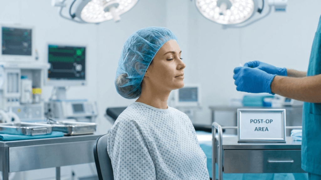 A medium shot, eye-level photograph set in a brightly lit, sterile operating room. A Caucasian female patient with a serene expression, closed eyes, and subtle, natural makeup sits in a hospital gown and a blue surgical cap. To her right, a medical professional wearing teal scrubs and blue latex gloves is seen from the side, holding a small medical instrument. The background features blurred high-tech medical monitoring equipment, surgical lights, and a metal cart with a 'POST-OP AREA' sign. The lighting is cool, clinical, and even, highlighting the clean, professional medical environment.