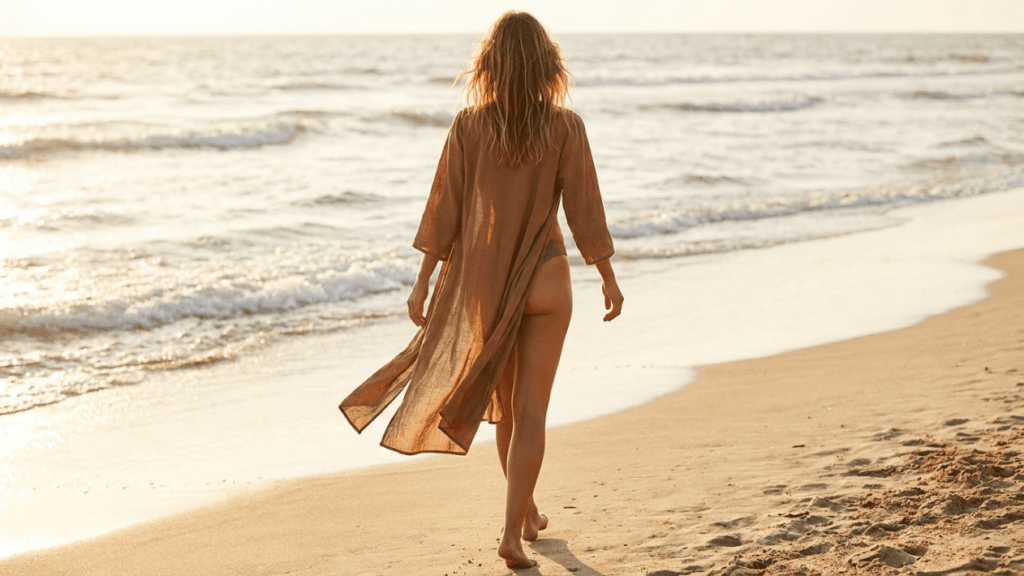 A woman with long, wavy, sun-kissed hair is viewed from behind, walking away along a sandy beach at sunset. The soft, warm golden hour lighting illuminates the scene, creating a gentle glow. She has a slender, feminine build and is wearing a sheer, flowy, brownish-tan long-sleeved cover-up over a bikini bottom. Her natural appearance includes no visible makeup, focusing on a relaxed, beachy aesthetic. The camera angle is at eye level, capturing her from a rear-facing perspective as she walks towards the calm ocean waves, which lap gently against the shore in the background.