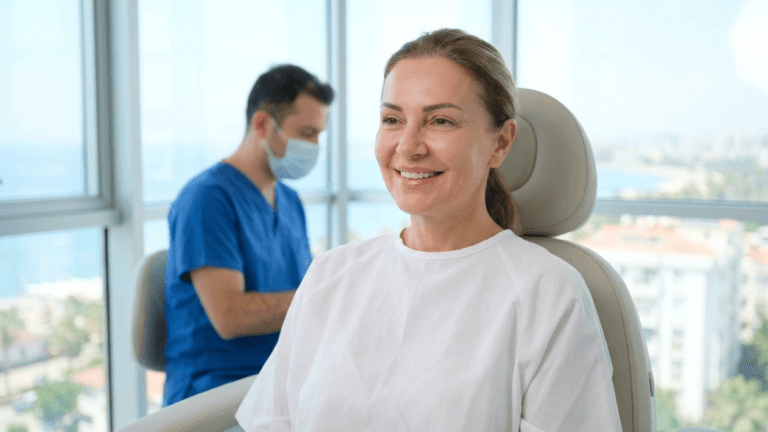 A middle-aged woman with a fair complexion and light brown hair tied back, wearing a simple white clinical top, sits smiling in a modern medical chair. She has light, natural makeup and a serene expression. Behind her, slightly out of focus, a male healthcare professional in a blue surgical scrub set and a protective face mask is visible. The scene is captured from a straight-on eye-level angle, bathed in bright, natural daylight streaming through large windows that reveal a blurred coastal cityscape in the background. The anatomy of the woman is relaxed, showing a calm demeanor in a professional clinic setting.