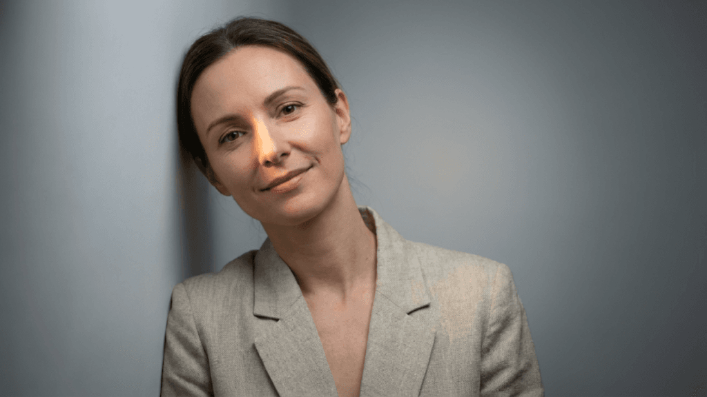 A professional portrait of a fair-skinned woman, facing the camera with a gentle smile and leaning against a neutral grey wall. The lighting is soft and directional, highlighting the side of her face. She has natural-looking makeup, shoulder-length brown hair pulled back, and is wearing a textured beige blazer. The shot is captured from a slightly elevated eye-level angle with a shallow depth of field, blurring the background.
