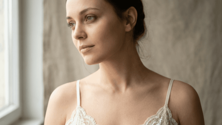 A portrait of a young woman with a feminine appearance, captured in a soft, natural side-angle shot. She has a serene, thoughtful expression, minimal makeup emphasizing a natural look, and her hair is tied back in a loose bun. She is wearing a delicate, white lace-trimmed camisole. The lighting is soft and diffused, coming from a nearby window, highlighting her facial features and slender neckline. The background consists of a neutral-toned, textured wall, creating a clean and minimalist aesthetic.