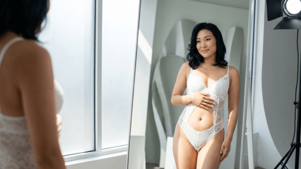 A medium shot of an East Asian woman with shoulder-length black hair looking at her reflection in a large floor mirror. She is feminine, smiling gently, and wearing a white lace bodysuit. Her anatomy is fit and toned, with her hand resting lightly on her abdomen. The lighting is soft and natural, coming from a large window on the left, complemented by a professional studio light on a tripod stand on the right. The camera angle is eye-level, capturing the subject and her reflection simultaneously in the mirror. The background is a bright, clean, modern studio space with abstract decorative shapes. Her makeup is natural and subtle.