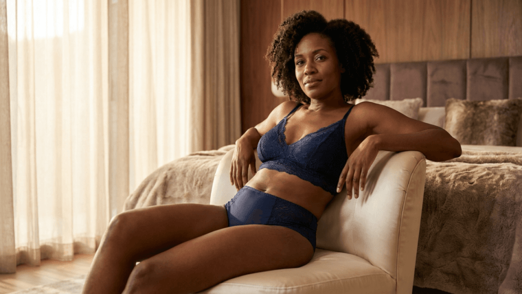 A confident Black woman with curly hair, wearing a navy blue lace bralette and matching high-waisted briefs, sits relaxed on a cream-colored armchair in a cozy, warm-toned bedroom. The lighting is soft and natural, coming from a window behind sheer curtains, creating a serene atmosphere. The shot is taken from a slightly elevated eye-level angle, focusing on her upper body and posture. She has subtle, natural-looking makeup, and her expression is calm and composed. The background features a plush bed with textured bedding, enhancing the intimate and comfortable setting.