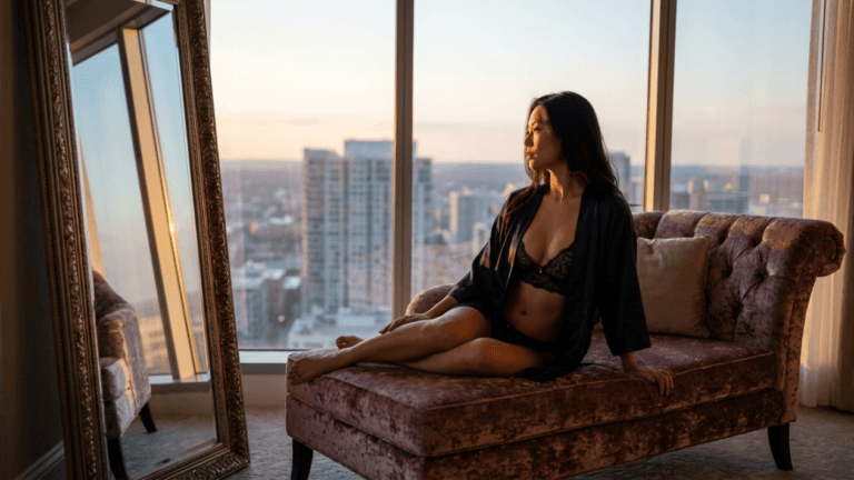 A beautiful woman with long dark hair, wearing a black lace bra set and a silk robe, reclines gracefully on a tufted velvet chaise lounge in a luxury high-rise apartment. The camera angle is at eye-level, capturing her profile as she gazes out of floor-to-ceiling windows toward a glowing, sunlit city skyline at sunset. The soft, golden ambient light illuminates her natural facial features and slender physique. To her side stands a tall, ornate gold-framed mirror, reflecting the opulent interior and the cityscape, adding depth to the warm, elegant atmosphere.