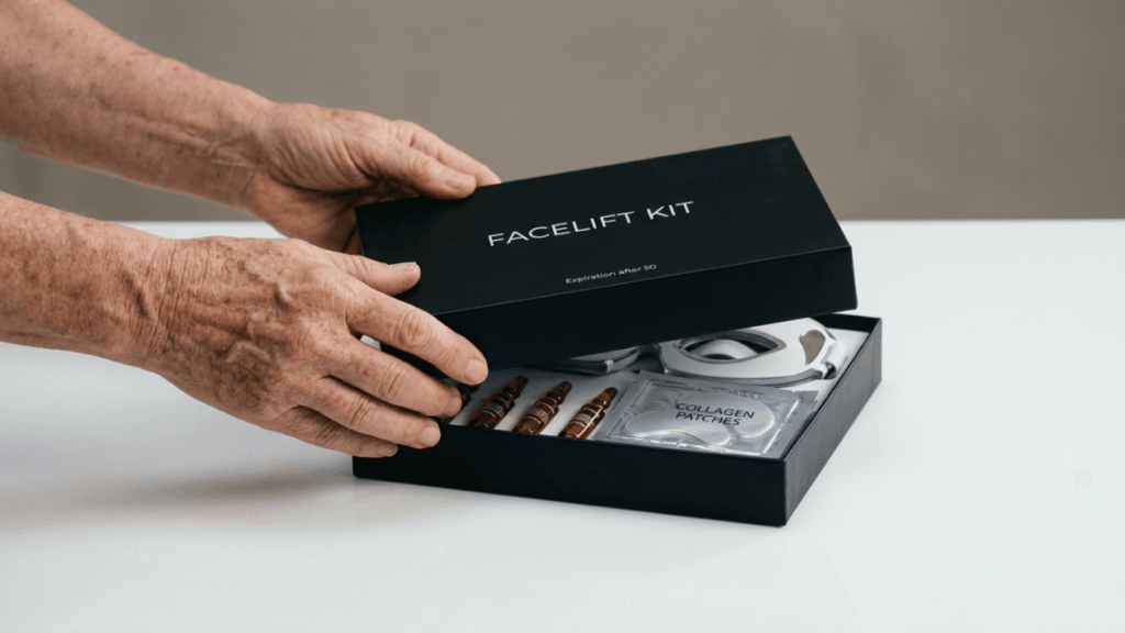 A pair of mature, wrinkled hands gently lifting the lid off a sleek black 'FACELIFT KIT' box on a white surface. The box contains small amber vials, a white facial device, and a silver 'COLLAGEN PATCHES' packet. The lighting is soft and neutral, the camera angle is at eye level, and the background is a plain, light grey studio setting. No makeup or clothing of the person is visible, focusing entirely on the hands and the product packaging.