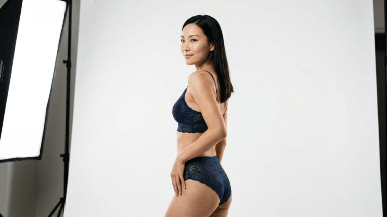 A studio portrait of a fit East Asian woman standing in profile, looking towards the camera with a gentle smile. She is wearing a dark blue lace bralette and matching high-waisted lace panties. The lighting is bright and professional, coming from a softbox on the left, casting soft shadows. She has straight, dark hair and natural, subtle makeup. The background is a clean, minimalist, seamless white studio backdrop, and the camera angle is at eye level, capturing a clear view of her upper body and posture.