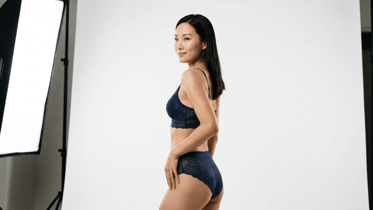A studio portrait of a fit East Asian woman standing in profile, looking towards the camera with a gentle smile. She is wearing a dark blue lace bralette and matching high-waisted lace panties. The lighting is bright and professional, coming from a softbox on the left, casting soft shadows. She has straight, dark hair and natural, subtle makeup. The background is a clean, minimalist, seamless white studio backdrop, and the camera angle is at eye level, capturing a clear view of her upper body and posture.