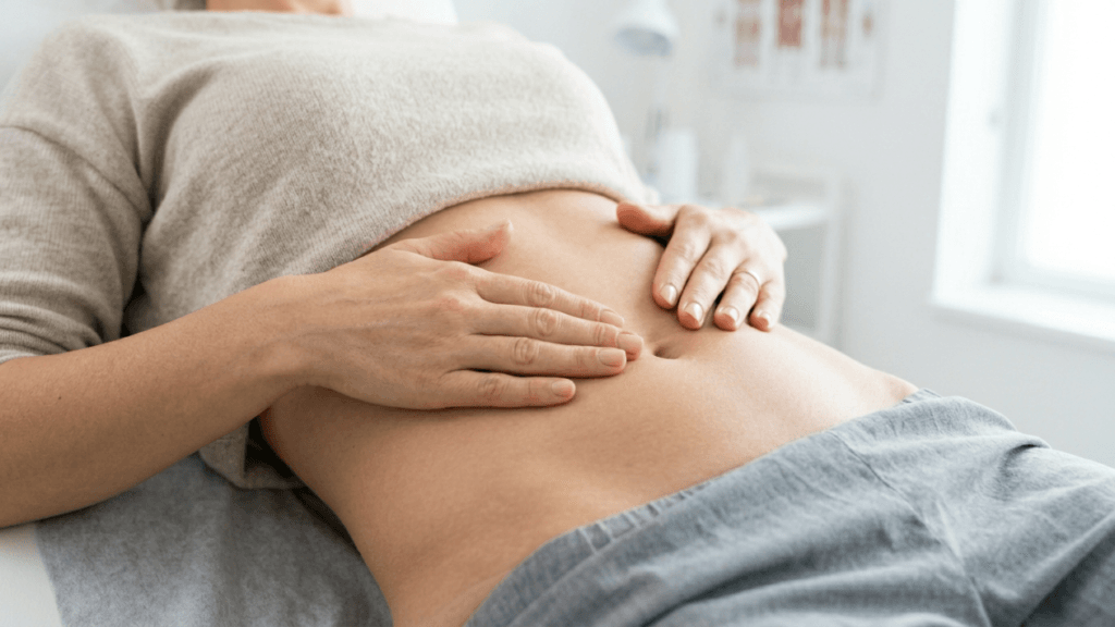 A close-up, high-angle shot of a female patient lying on an examination table in a bright, clinical setting. Her hands are gently resting on her exposed abdomen. The lighting is soft and natural, emphasizing the calm medical environment. She is wearing a casual beige top and grey sweatpants. The focus is on the midsection anatomy, with a blurred medical office background featuring white walls and a window. No makeup is visible as the focus is solely on the torso.
