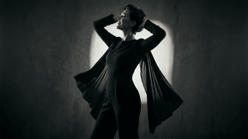 A cinematic, dramatic portrait of an elegant woman with short, styled hair, captured from a side-profile angle against a dark, textured grey background. She is illuminated by a soft, circular spotlight behind her head, creating a subtle halo effect. She wears a sophisticated, long-sleeved black jumpsuit featuring flowing, cape-like fabric attached to the sleeves that gracefully extends outwards as she raises her arms behind her head. Her expression is serene and confident, with soft, natural-toned makeup highlighting her features. The lighting emphasizes her silhouette and the flowing movement of the fabric, conveying a sense of artistic poise and modern sophistication.