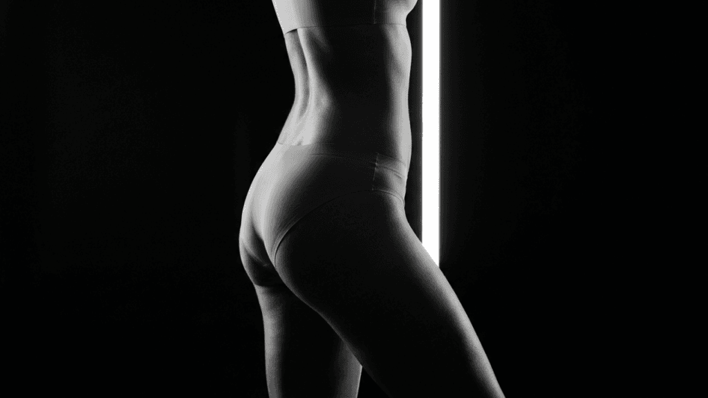 A professional black and white studio portrait of a woman's lower torso and hips, captured from a side profile angle. The lighting is high-contrast, featuring a vertical glowing light strip in the background that highlights the contours of her body. The woman has a feminine, fit anatomy and is wearing form-fitting seamless underwear. The shot focuses on body aesthetics, with a minimalist, clean aesthetic and deep black background. No makeup is visible as the image focuses solely on torso and body shape.