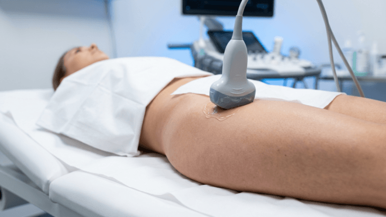 A female patient lying on a medical examination table undergoes a hip ultrasound procedure; the scene features bright clinical lighting from a side angle, with a medical professional applying an ultrasound probe to the patient's hip area covered in gel, against a blurred background of a diagnostic ultrasound machine.