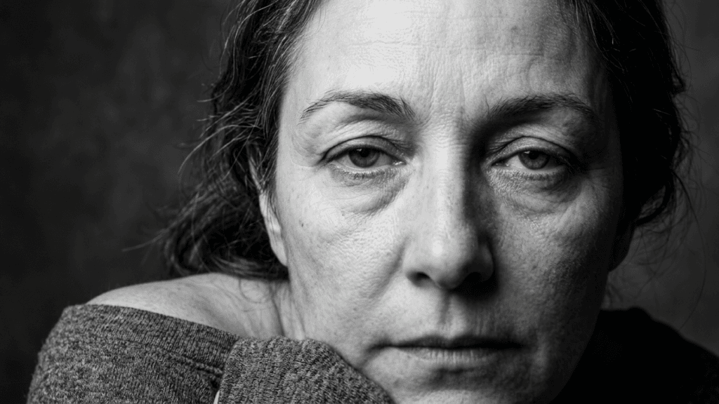 A close-up, black and white portrait of a middle-aged woman with a natural, unadorned appearance. She has a serious, contemplative expression and is looking directly into the camera. The lighting is soft and directional, highlighting the natural textures and fine lines of her skin, emphasizing an authentic and aged look. She wears a simple, casual grey top that rests off one shoulder. The camera angle is eye-level, creating an intimate connection with the viewer. Her face is framed by natural, slightly messy hair, and the background is a neutral, dark, and out-of-focus textured surface. No makeup is visible, focusing entirely on her natural features and anatomy.