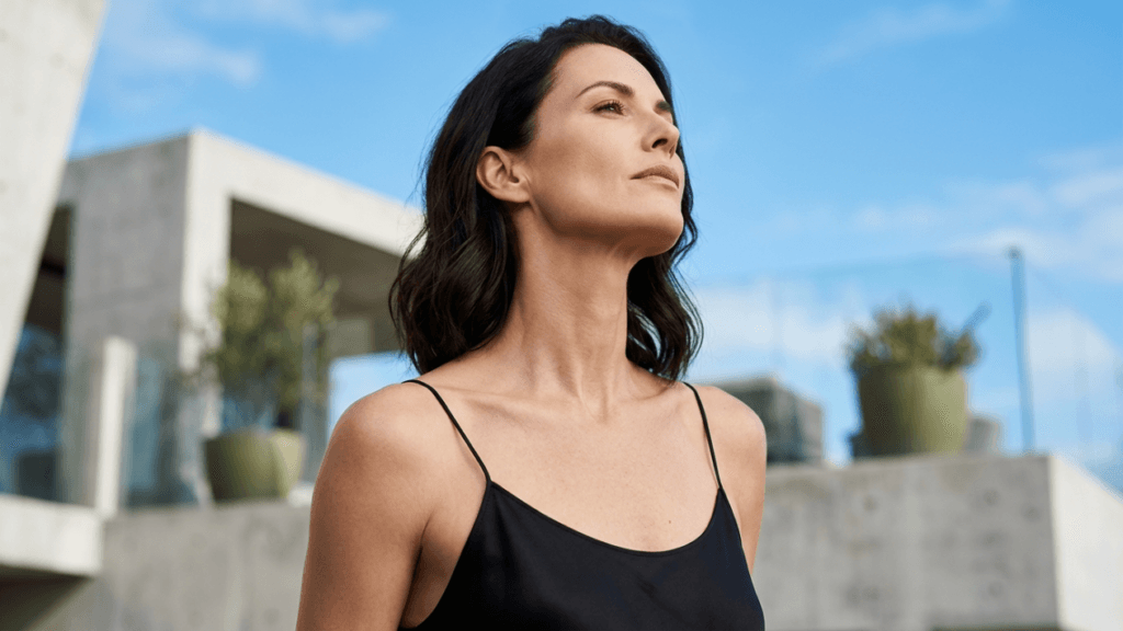 A professional portrait of a woman with long, dark wavy hair, captured from a medium low-angle perspective. She is facing slightly upwards and to the side, showcasing elegant facial anatomy and a refined jawline. She wears minimal, natural makeup and a simple black spaghetti-strap top. The lighting is bright and soft, typical of natural daylight, creating a clean aesthetic. The background features a modern, architectural structure with minimalist concrete elements and blurred green plants under a clear, bright blue sky, emphasizing a sophisticated and serene atmosphere.