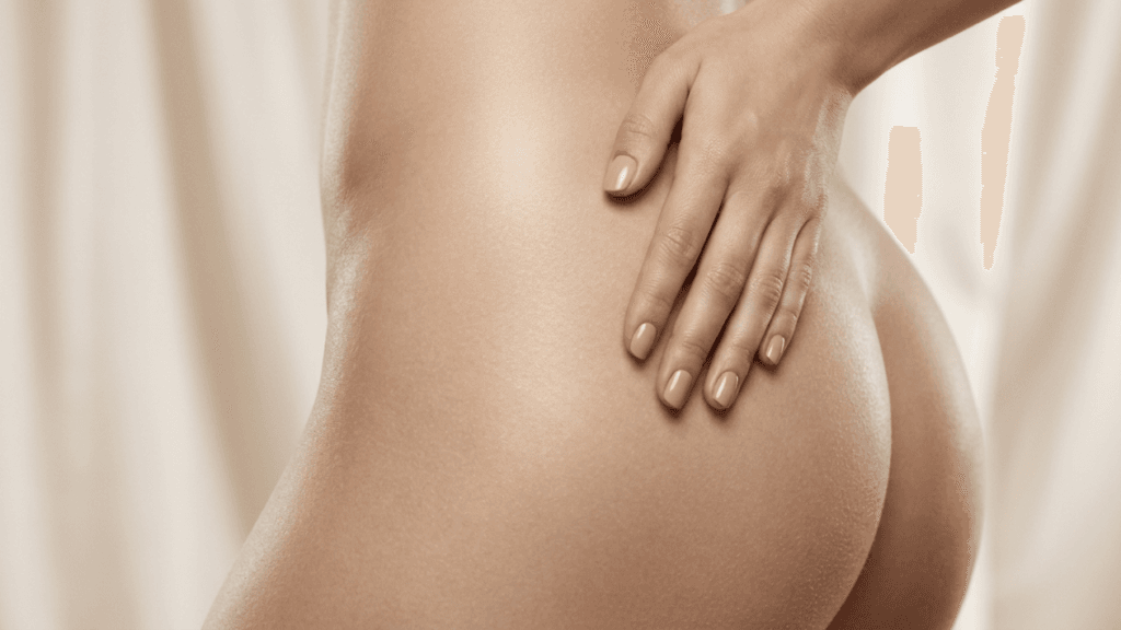 A close-up, studio-lit shot of a feminine physique, focusing on the hip and gluteal area. The skin is smooth with a subtle, healthy sheen. A hand with neatly manicured, nude-colored nails rests gently on the hip. The lighting is soft and even, highlighting the natural contours of the body. The background is a soft, out-of-focus fabric drape in a neutral, creamy tone, creating a clean and professional aesthetic. No clothing is visible.